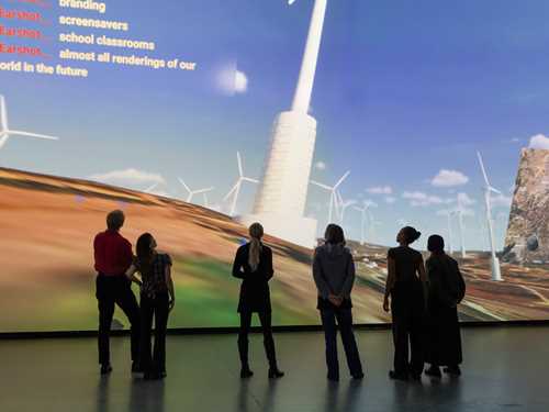 A group of people stand in a dark room in front of a wall covered by a projection showing a computer animation of wind turbines on the Golan Heights. Photograph