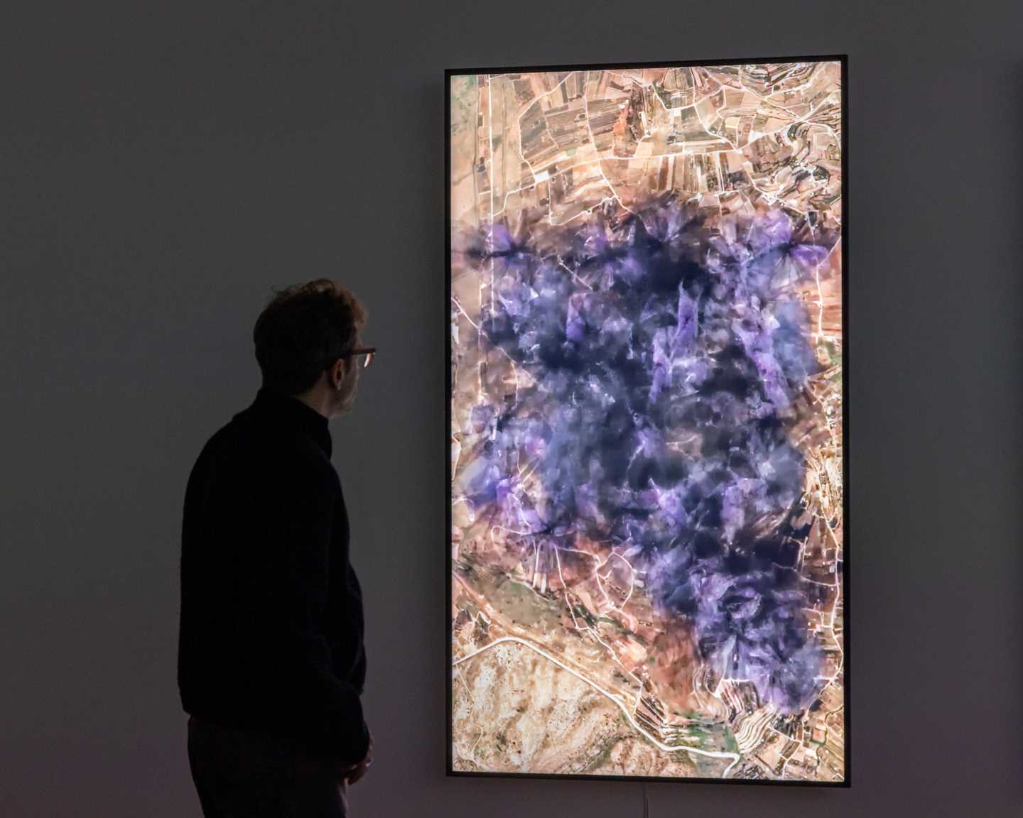 The artist Lawrence Abu Hamdan stands in a dark room, looking at a vertical screen showing a visualisation of the noise generated by 31 wind turbines on the Golan Heights. On the screen, the noise spreads across a vast area like purple and black crackling smoke. Photograph