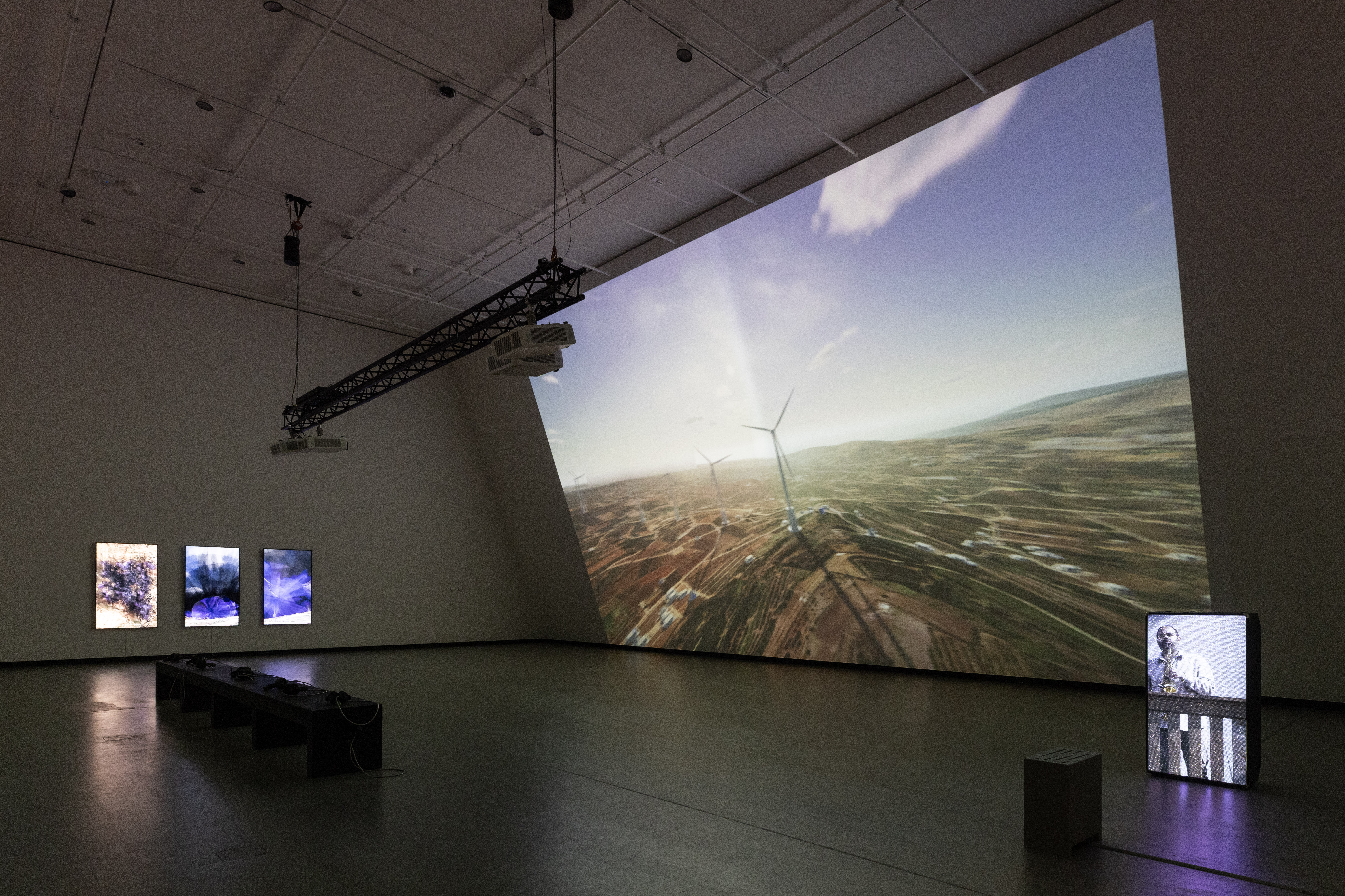 A dark exhibition room illuminated by a projection covering an entire wall. The projection shows a computer animation of wind turbines on the Golan Heights. In the middle of the room stands a black bench, and on the wall to the left of the image hang three vertical screens. On the far right of the image, a loudspeaker displays a projection of the saxophonist Amr Mdah playing from a balcony. Photograph