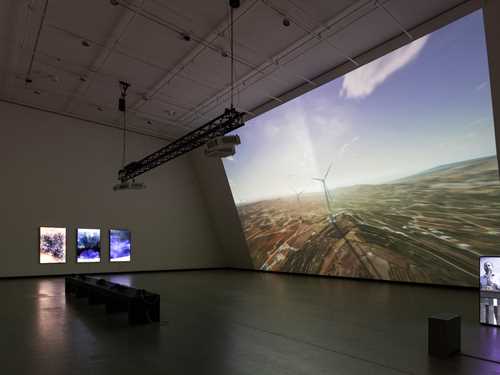 A dark exhibition room illuminated by a projection covering an entire wall. The projection shows a computer animation of wind turbines on the Golan Heights. In the middle of the room stands a black bench, and on the wall to the left of the image hang three vertical screens. On the far right of the image, a loudspeaker displays a projection of the saxophonist Amr Mdah playing from a balcony. Photograph