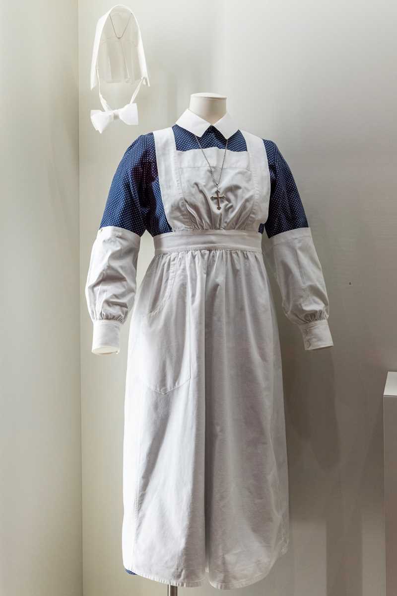 Deaconess uniform on a mannequin: white shirt, blue spotted skirt, white apron, detachable sleeves, black tights, and a white nurse’s cap with chin strap. The cap symbolises the completion of training.