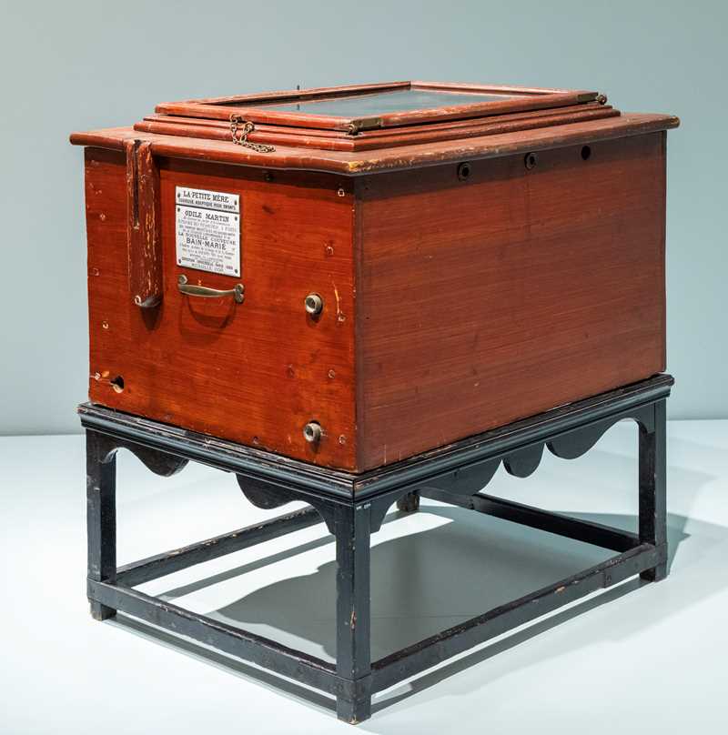 An incubator made of dark stained wood is mounted on a stand of black stained wood carved with decorative waves. The lid has a built-in glass plate that lets you look inside the box.