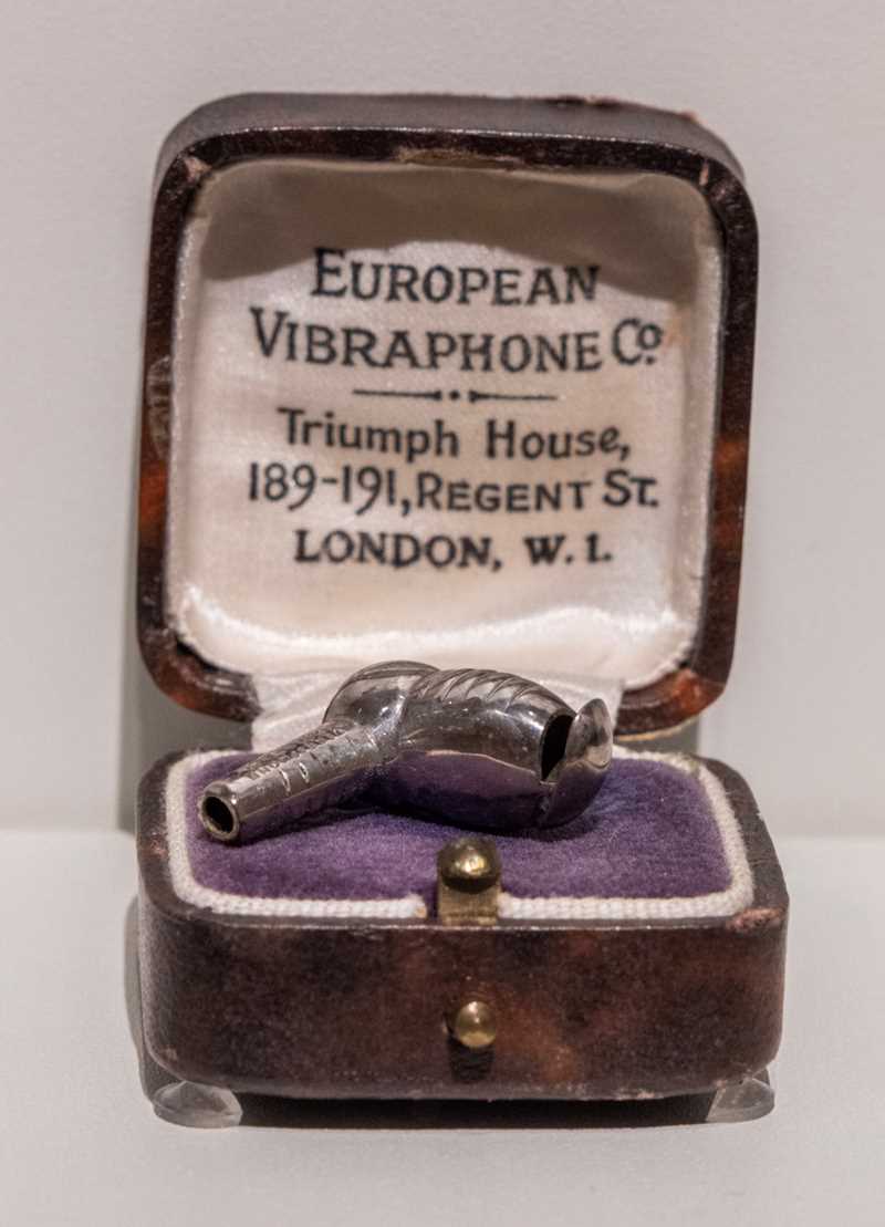 A small leather case, lined with a white silk fabric. In the base of the box is a purple velour cushion, which holds a silver hearing aid.