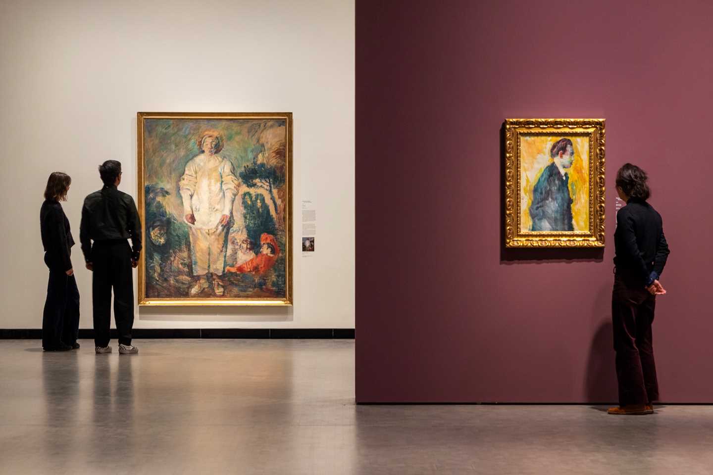 Image from an exhibition hall showing three people looking at two different works by Ludvig Karsten. Photograph.