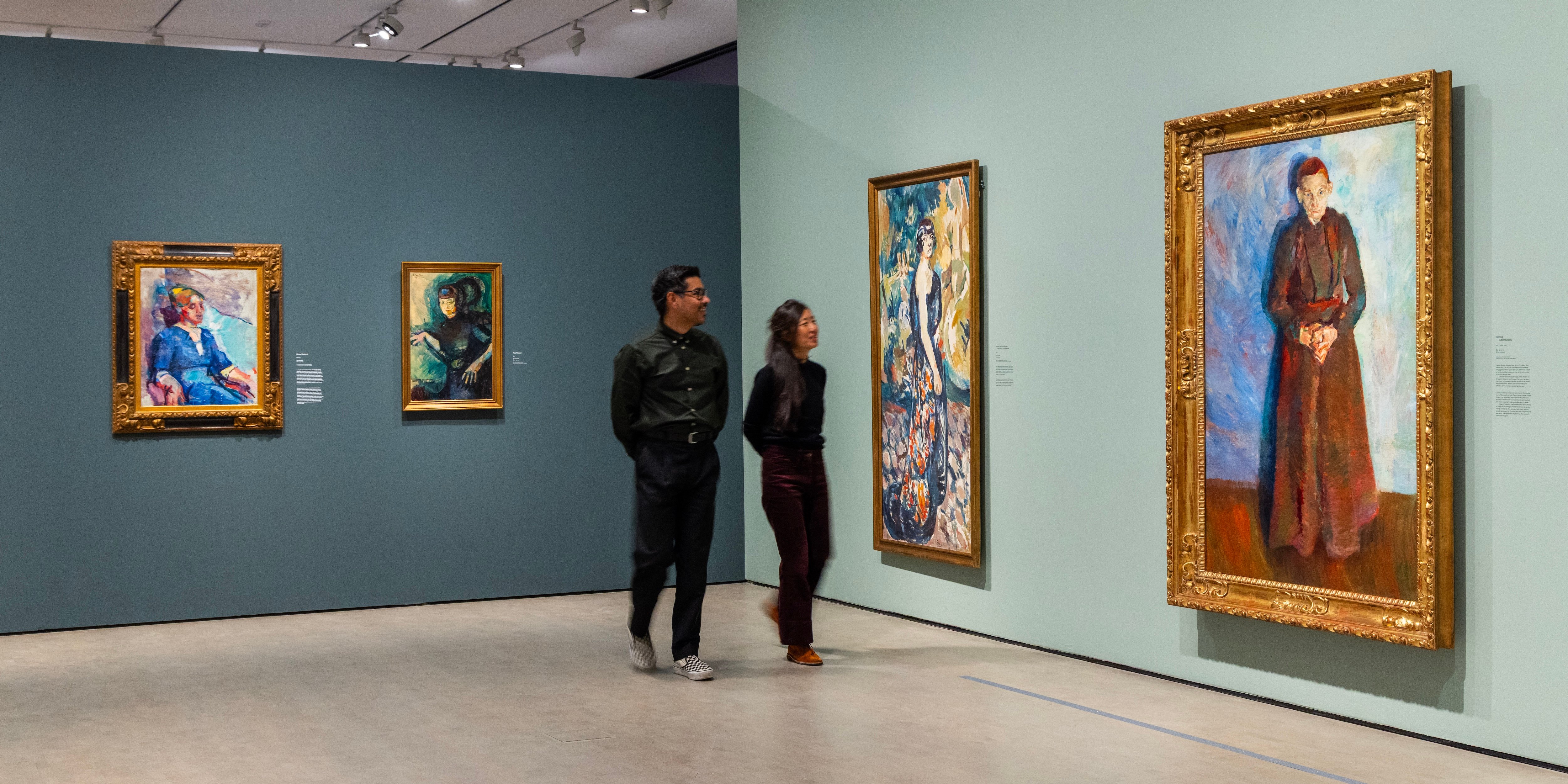 Two people walking through an exhibition, smiling as they look at paintings by Ludvig Karsten. Photograph.