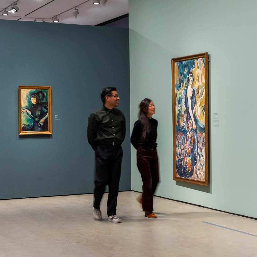 Two people walking through an exhibition, smiling as they look at paintings by Ludvig Karsten. Photograph.