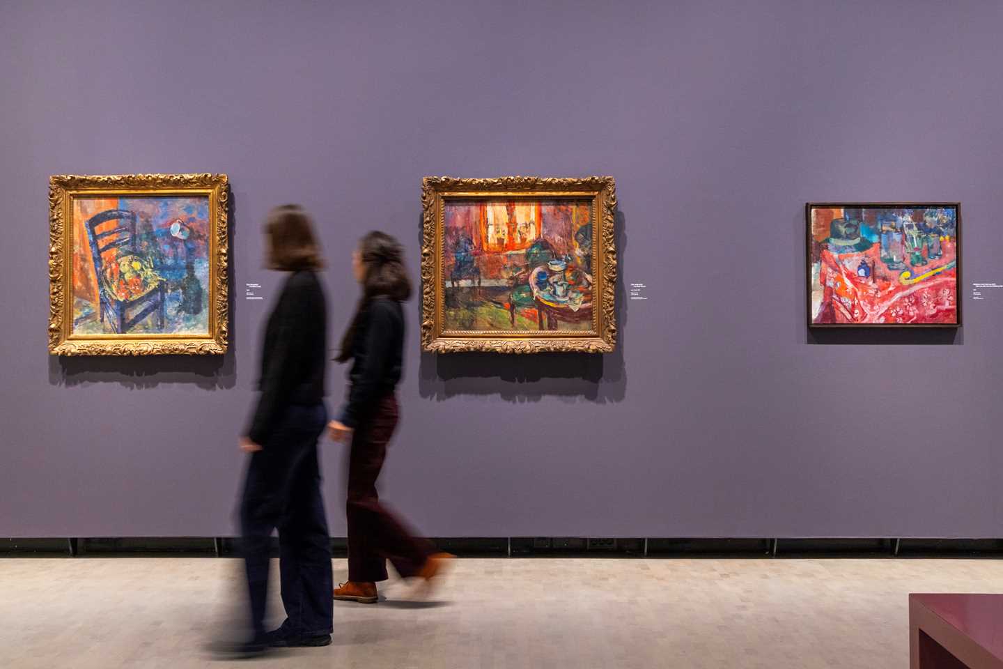Two women walking along a purple wall with three paintings by Ludvig Karsten. Photograph.