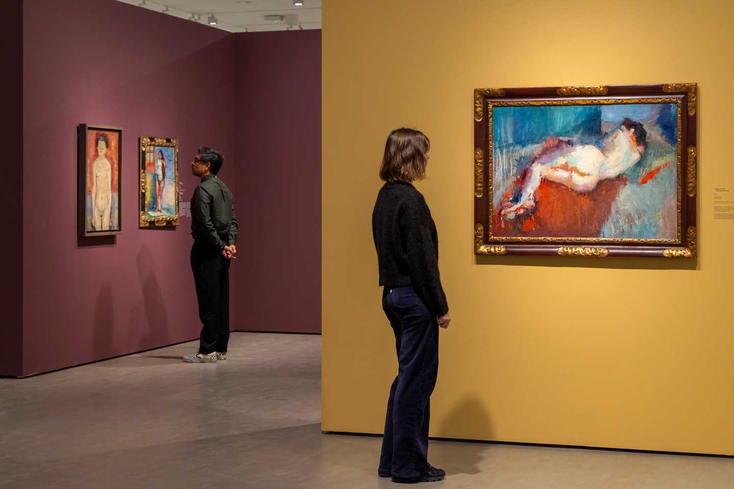 Two people looking at works by Ludvig Karsten. Closest to the camera, a woman is viewing the painting Reclining Nude. Photograph.