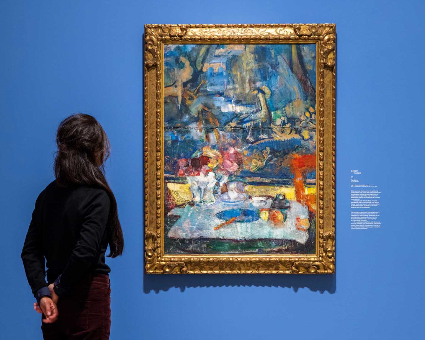 A woman looking at Gobelin by Ludvig Karsten, hanging on a blue wall in a large gold frame. Photograph.