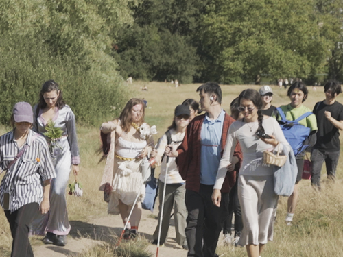 A group of people walking along a cart road in a landscape of forest and fields. It is a sunny summer day, and several of the people are using white canes. Photograph.