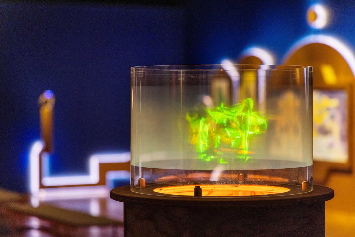 A hologram showing dancing green figures is in focus. Behind it, a blue exhibition room and another art installation are visible. Photograph.