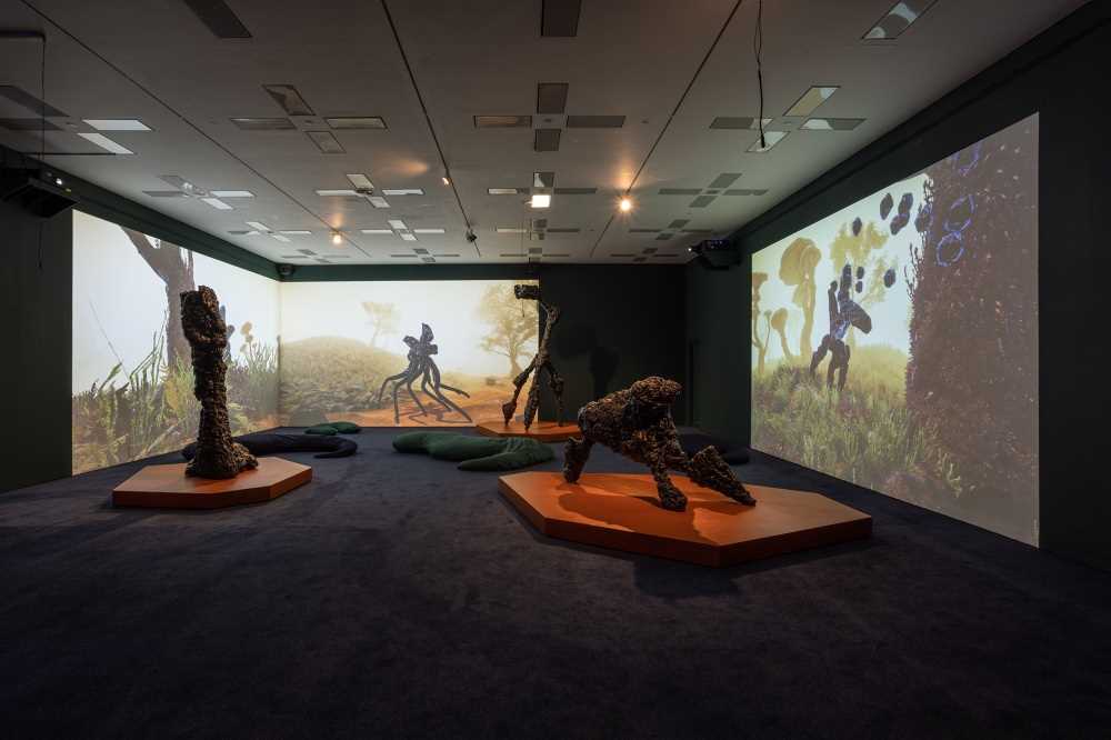 An exhibition room with projections on three walls and three sculptures standing on the floor alongside some elongated cushions. Photograph.