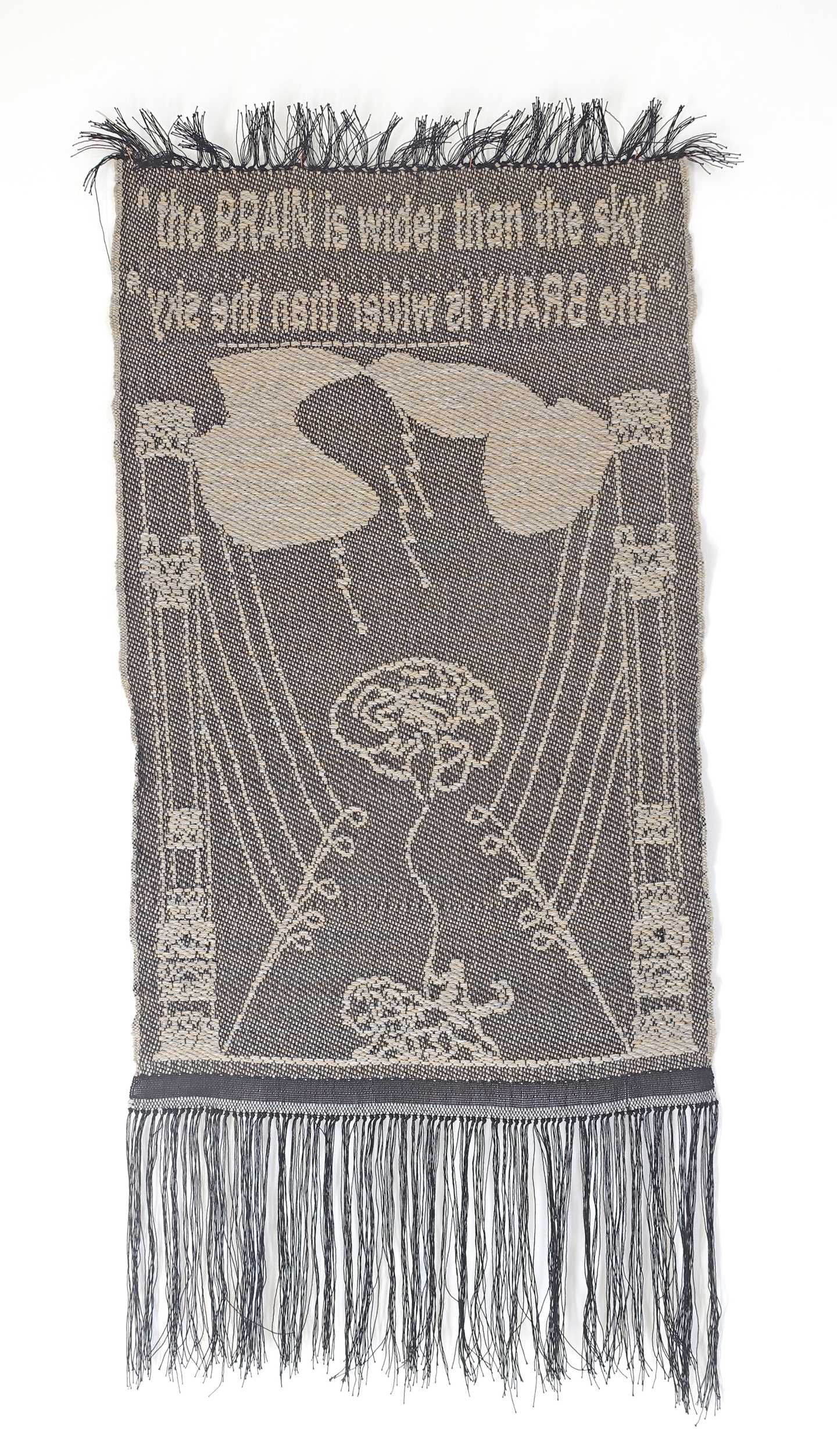A tapestry in shades of grey with the text ‘The BRAIN is wider than the sky.’ In the centre is a brain, and above it cloud-like shapes from which lightning strikes descend. Photograph.