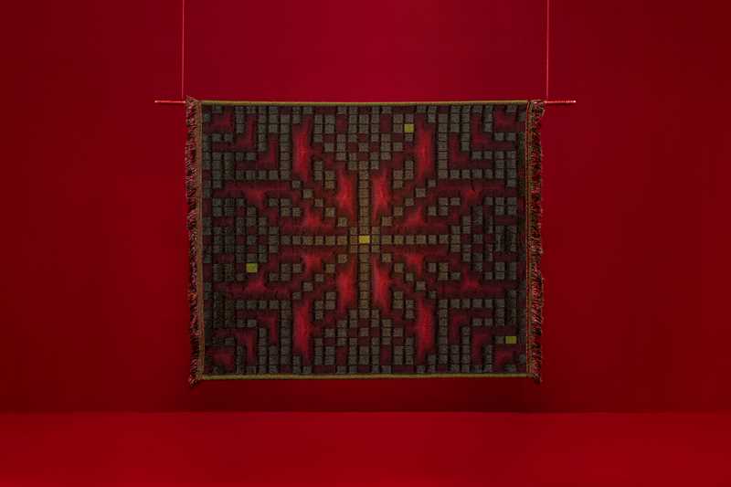 A square tapestry hangs against a red background. The pattern resembles the Selbu rose but is composed of small boxes that aesthetically resemble Tetris or similar video game blocks. Photograph.