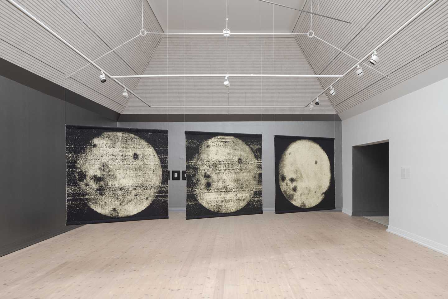 Three square tapestries of the moon hang side by side from the ceiling in a diagonal line in an exhibition room. Photograph.