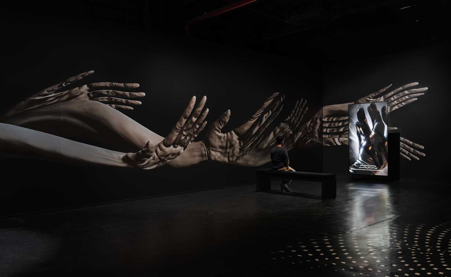 A dark exhibition room where animated hands move along the walls. In the centre of the room is a bench with a man sitting and watching a vertical screen. Photograph.