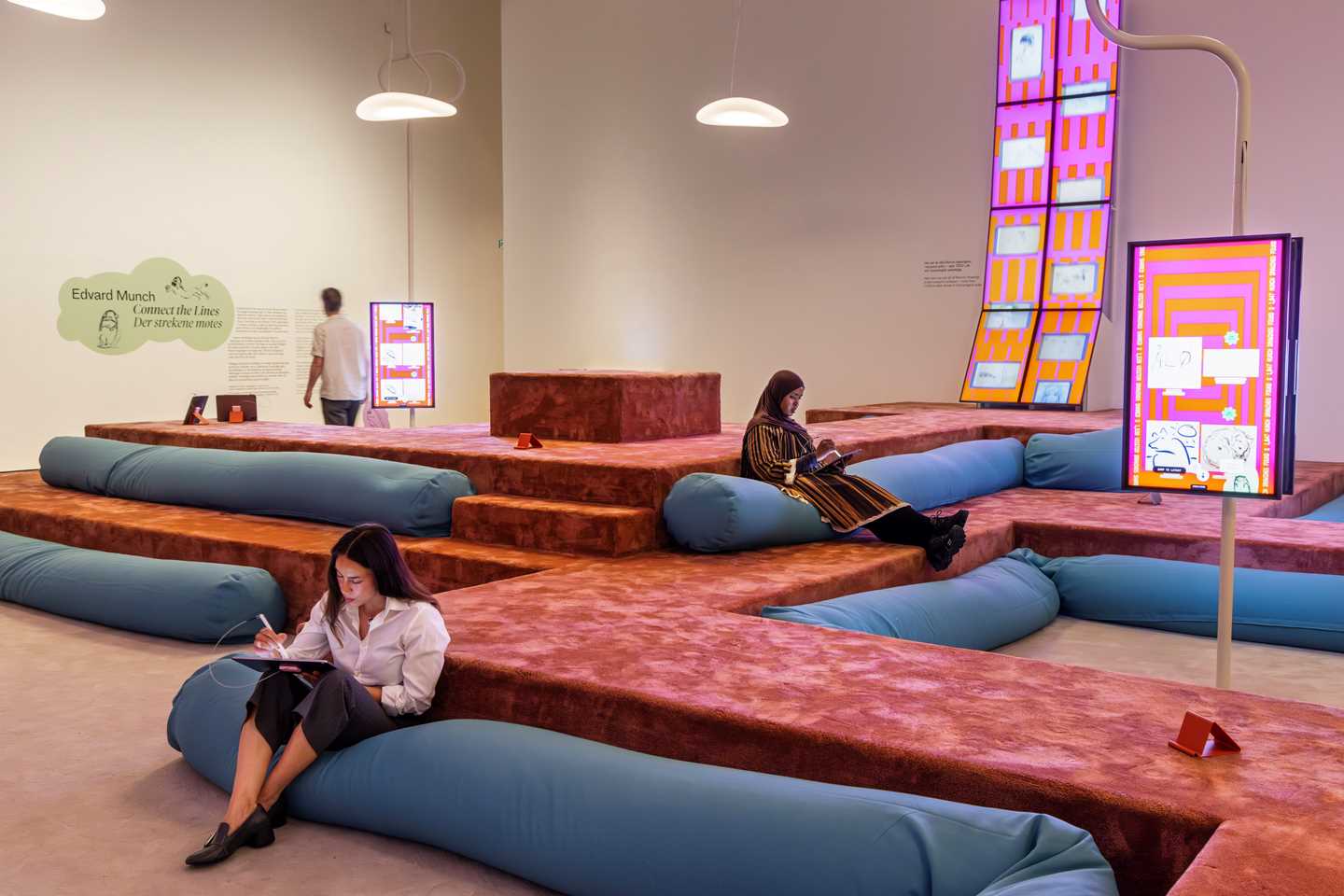Room filled with long blue beanbags arranged on a soft terracotta-coloured amphitheatre. Screens around the space display vivid pink and orange visuals of drawings by Edvard Munch and by visitors. Two people sit drawing on iPads, while a third stands reading wall text. Photograph.