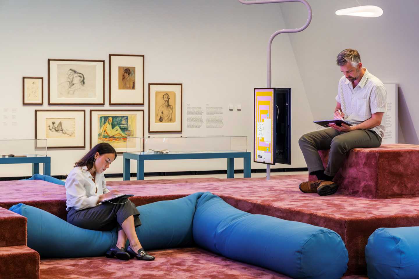 Woman seated on a beanbag drawing on an iPad, while a man sits atop an amphitheatre drawing on an iPad. Original drawings by Edvard Munch hang on the wall in the background. Photograph.