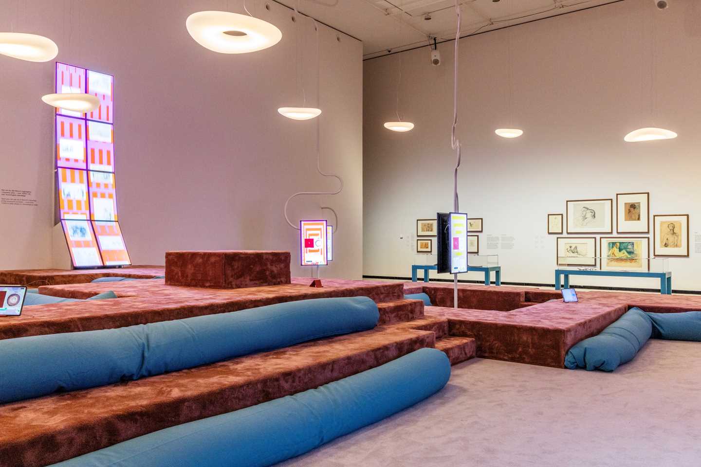 Overview shot of the gallery showing the amphitheatre with beanbag cushions, screens displaying drawings by Edvard Munch and visitors, as well as ceiling lights resembling clouds. Photograph.