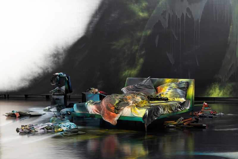 An unmade bed on a black-painted floor covered in spray paint. The wall behind is also spray-painted black, and numerous objects, likewise spray-painted, are arranged on the floor around the bed. Photograph.