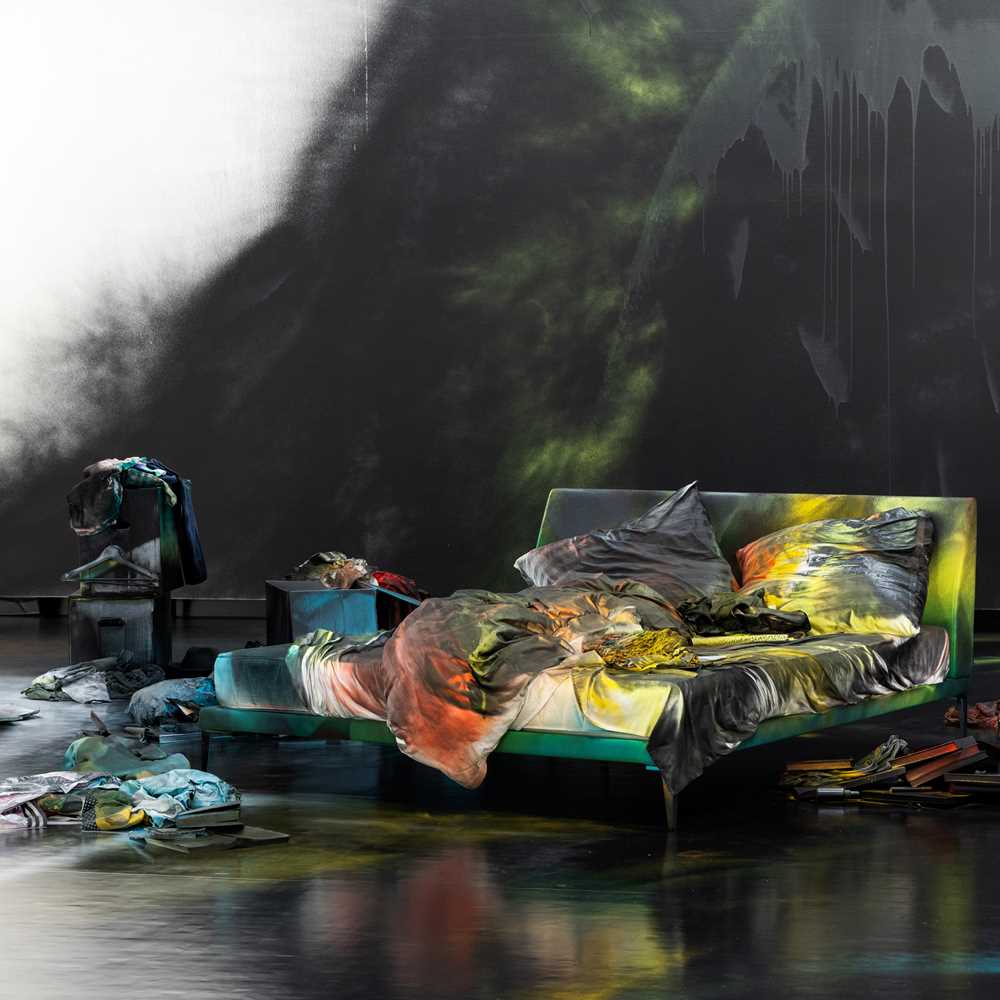 An unmade bed on a black-painted floor covered in spray paint. The wall behind is also spray-painted black, and numerous objects, likewise spray-painted, are arranged on the floor around the bed. Photograph.