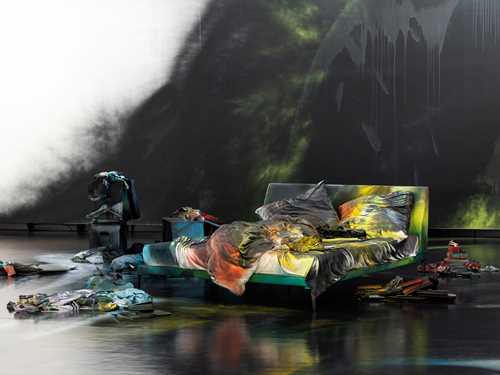 An unmade bed on a black-painted floor covered in spray paint. The wall behind is also spray-painted black, and numerous objects, likewise spray-painted, are arranged on the floor around the bed. Photograph.