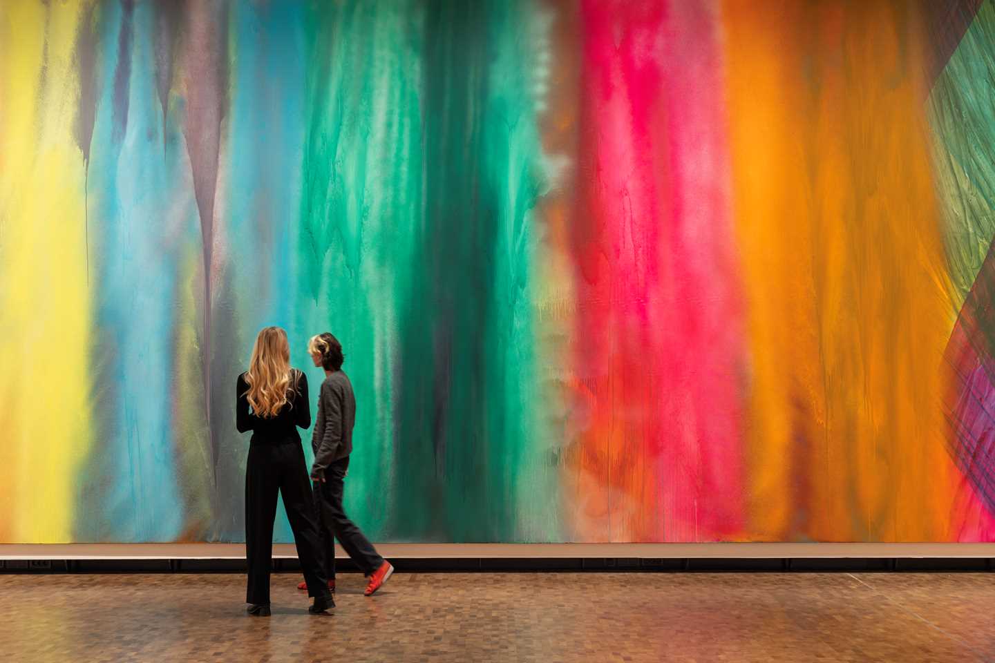 Two women walk past a vast painting that covers the entire wall behind them. Photograph.