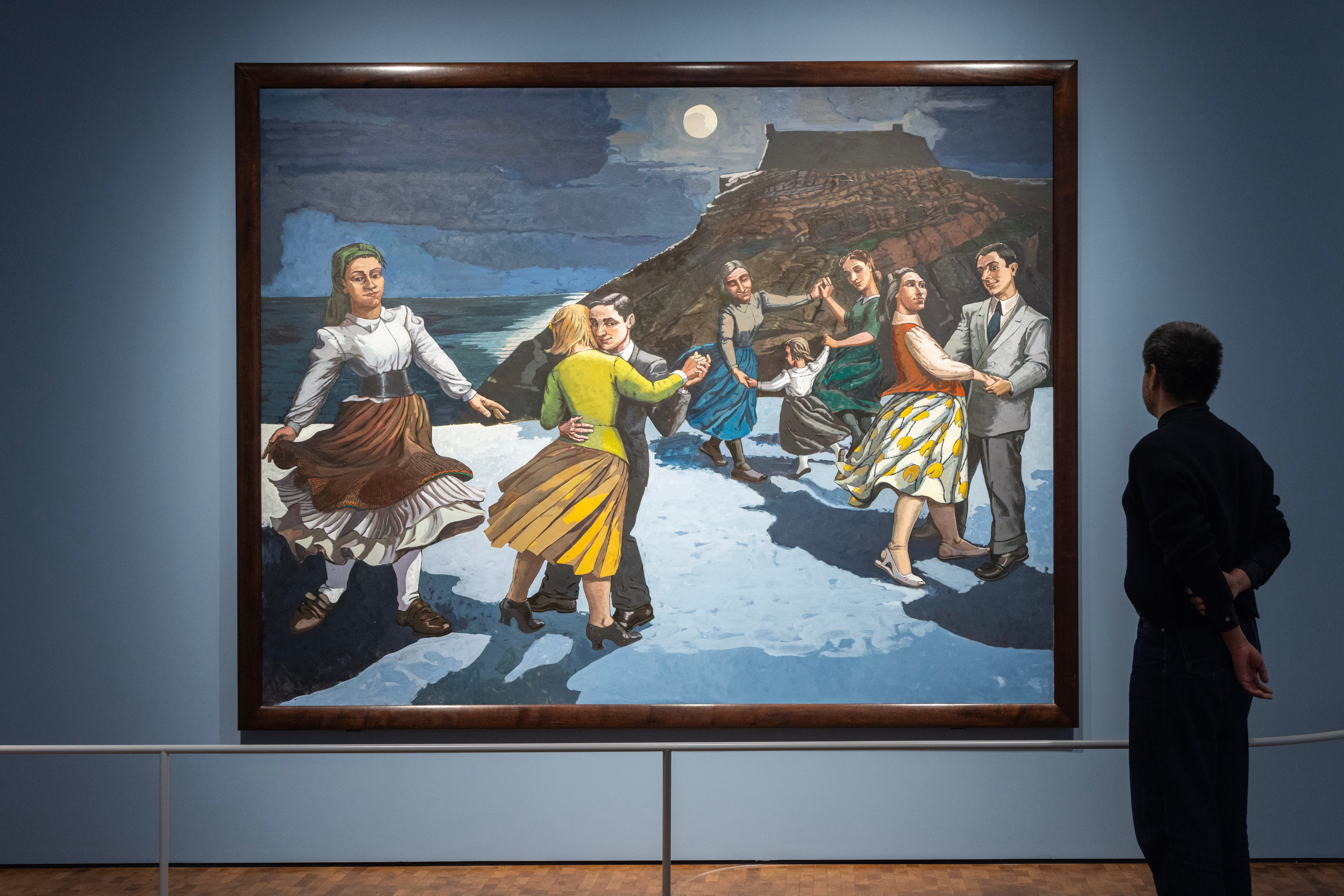 A man stands with his back to the camera, looking at the painting The Dance by Paula Rego. The painting shows a beach with a mountain in the background and a dark sky with a glowing moon. On the beach, several couples are dancing, along with a woman dancing alone. Photograph.