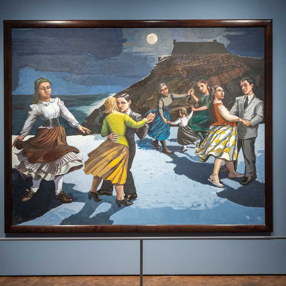 A man stands with his back to the camera, looking at the painting The Dance by Paula Rego. The painting shows a beach with a mountain in the background and a dark sky with a glowing moon. On the beach, several couples are dancing, along with a woman dancing alone. Photograph.