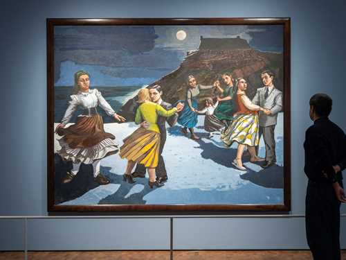 A man stands with his back to the camera, looking at the painting The Dance by Paula Rego. The painting shows a beach with a mountain in the background and a dark sky with a glowing moon. On the beach, several couples are dancing, along with a woman dancing alone. Photograph.