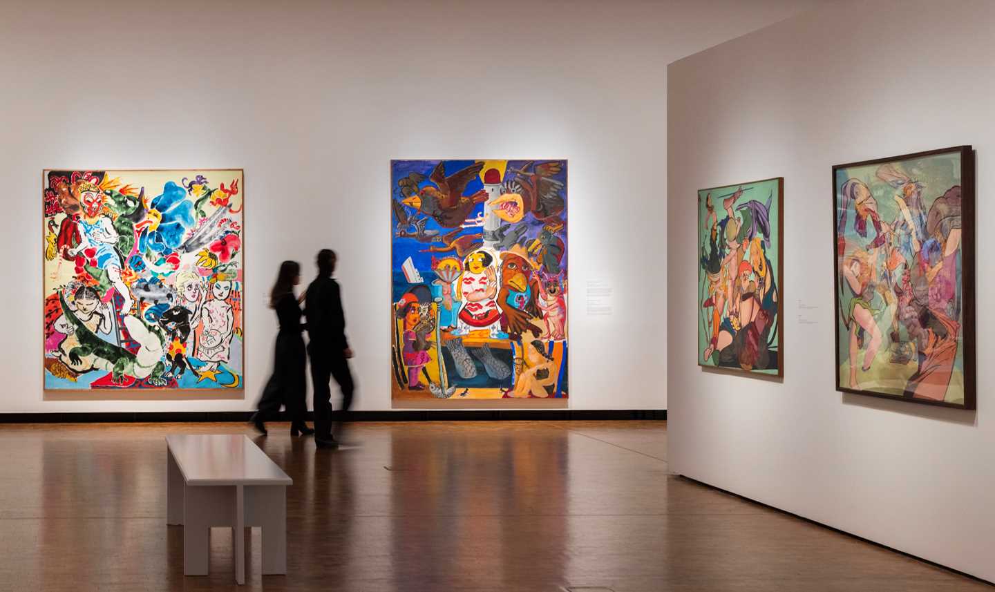 A view from an exhibition gallery showing works by Paula Rego. In this image, four colourful works hang on white walls, while two people walk through the space. Photograph.