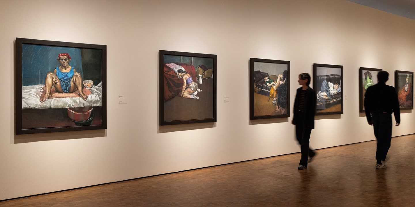 A view from an exhibition gallery showing works by Paula Rego. In this image, six works from her abortion series hang on white walls, while two people walk through the space. Photograph.