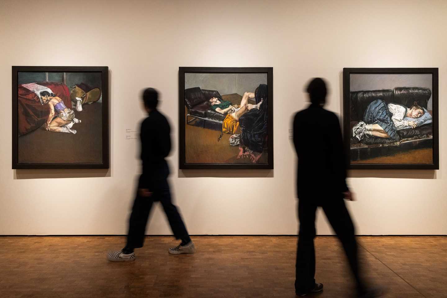 A view from an exhibition gallery showing works by Paula Rego. In this image, three works from her abortion series hang on white walls, while two people walk through the space. Photograph.