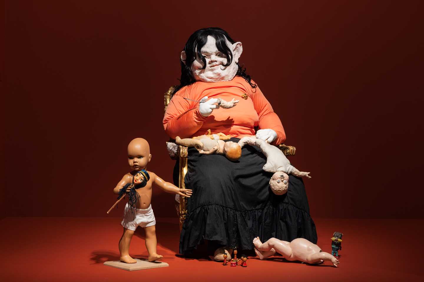 A sculpture of a seated, human-like female figure surrounded by fragmented childlike forms arranged around her. One intact childlike figure stands on the floor beside her, untouched. Photograph