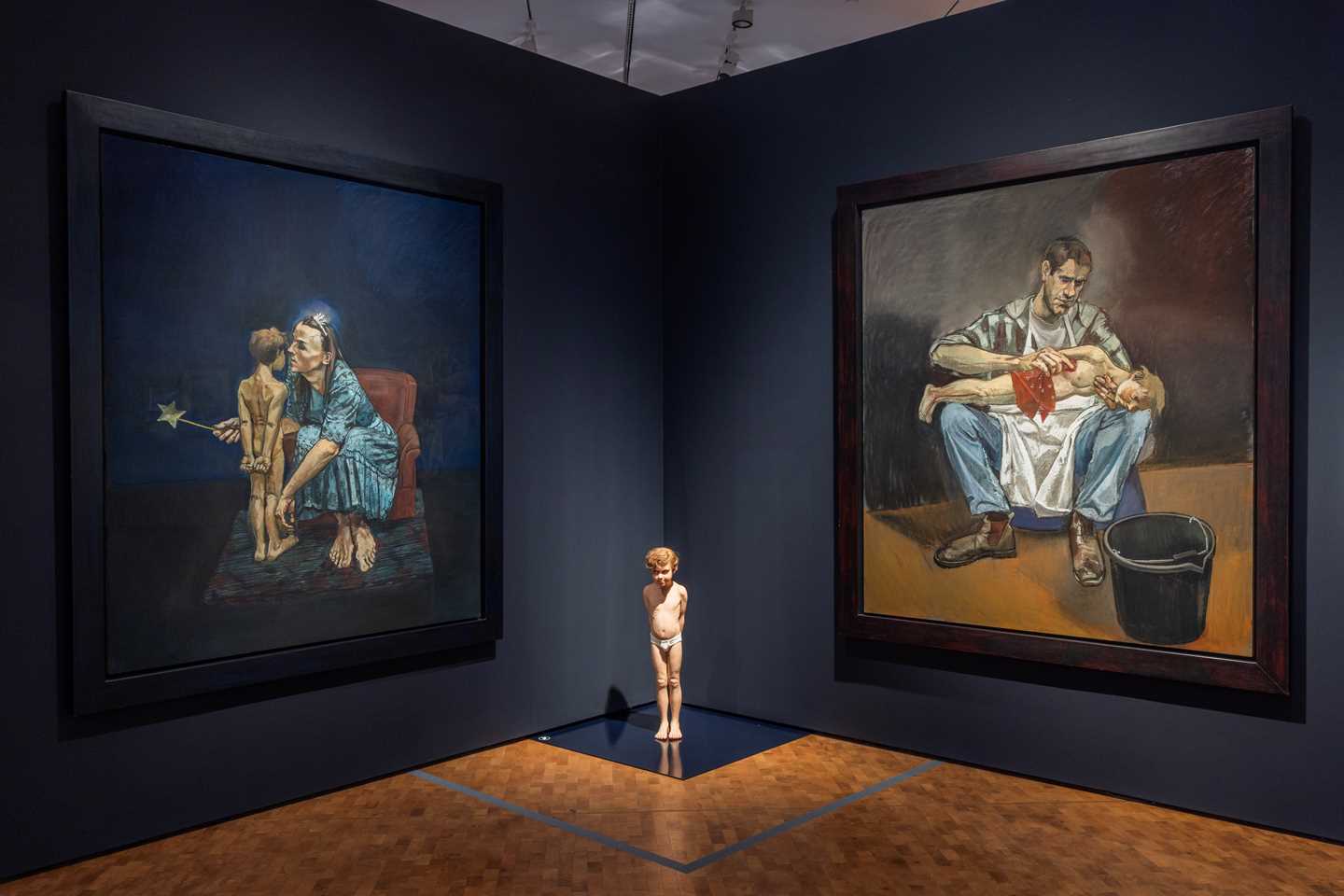 Against a dark blue wall hang two large works: one showing a woman with a small boy, and the other a man with the same boy. On the floor between the pastels stands a sculpture of the boy. Photograph.