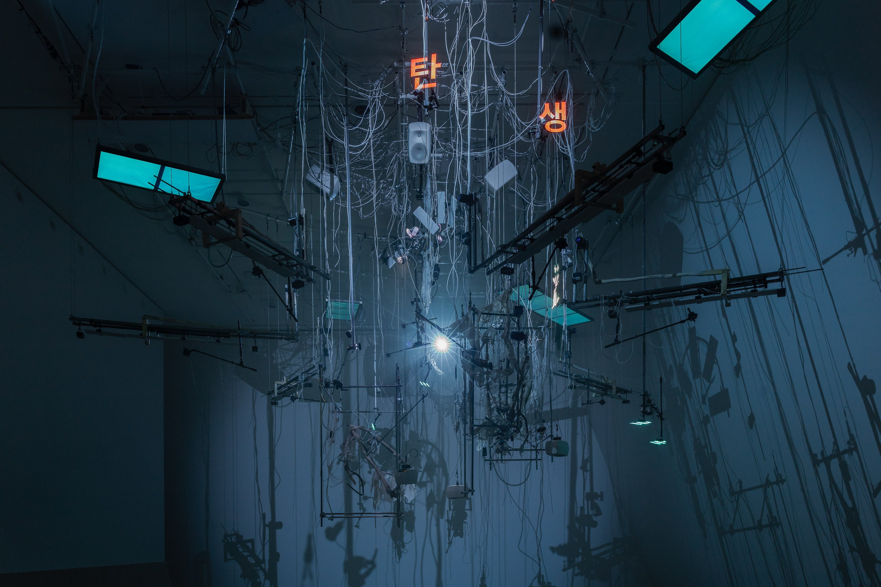 An installation hangs from the ceiling in a dark room illuminated by blue light. The installation includes numerous wires, instrument strings, light screens, and keyboards. Photograph.