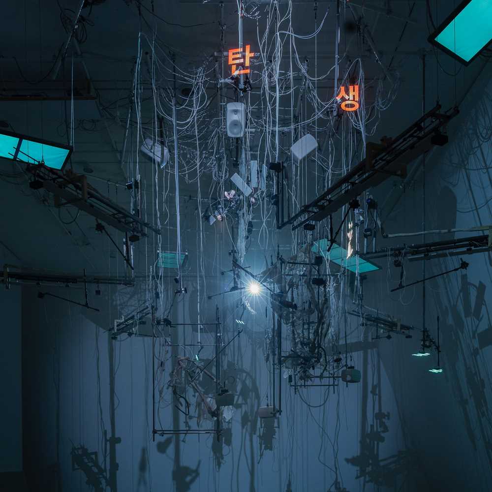 An installation hangs from the ceiling in a dark room illuminated by blue light. The installation includes numerous wires, instrument strings, light screens, and keyboards. Photograph.