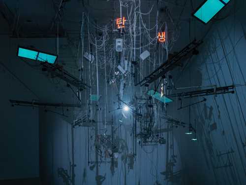 An installation hangs from the ceiling in a dark room illuminated by blue light. The installation includes numerous wires, instrument strings, light screens, and keyboards. Photograph.