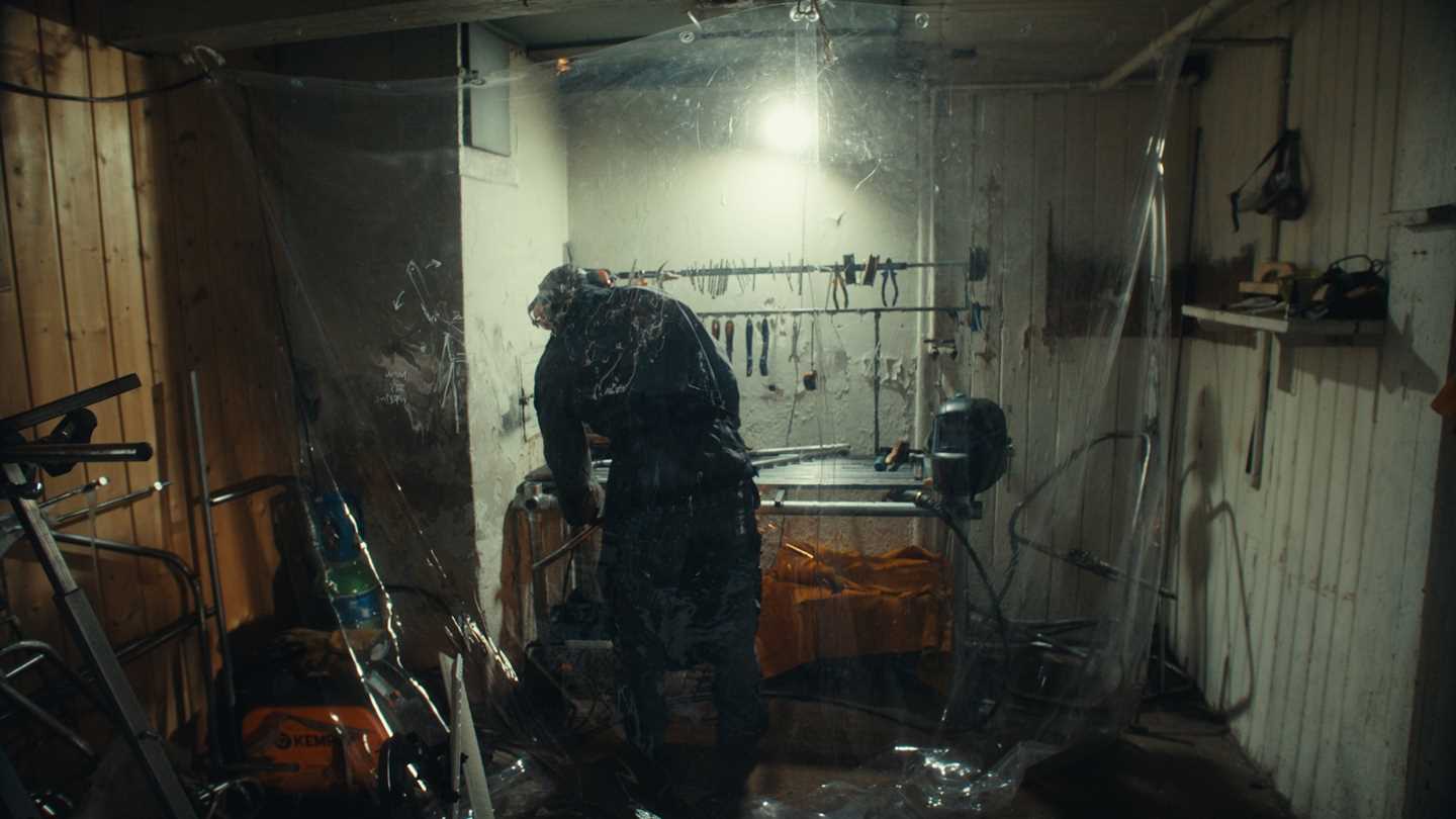 A man in overalls stands with his back to the camera in a workshop, with transparent plastic hanging from the ceiling. Photograph.