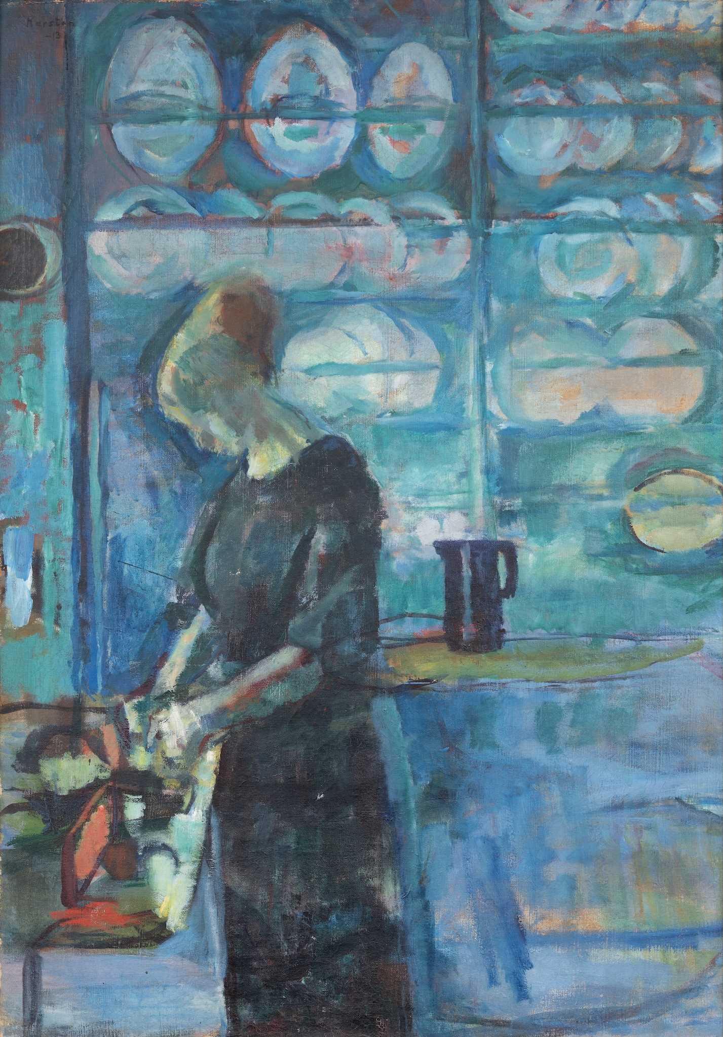A woman with her hair tied up and wearing a dark dress stands by a kitchen counter, seen in profile. Behind her is a wall covered with shelves holding upright plates and dishes. In the background there is also a table with a jug on it. Painting.