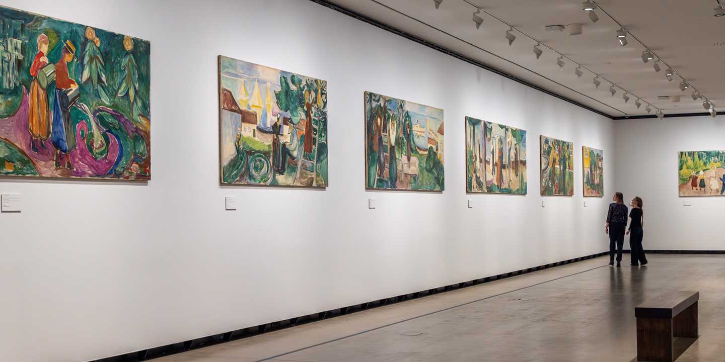 Two women look at works from the Freia Frieze hanging along a long white wall. Photograph.
