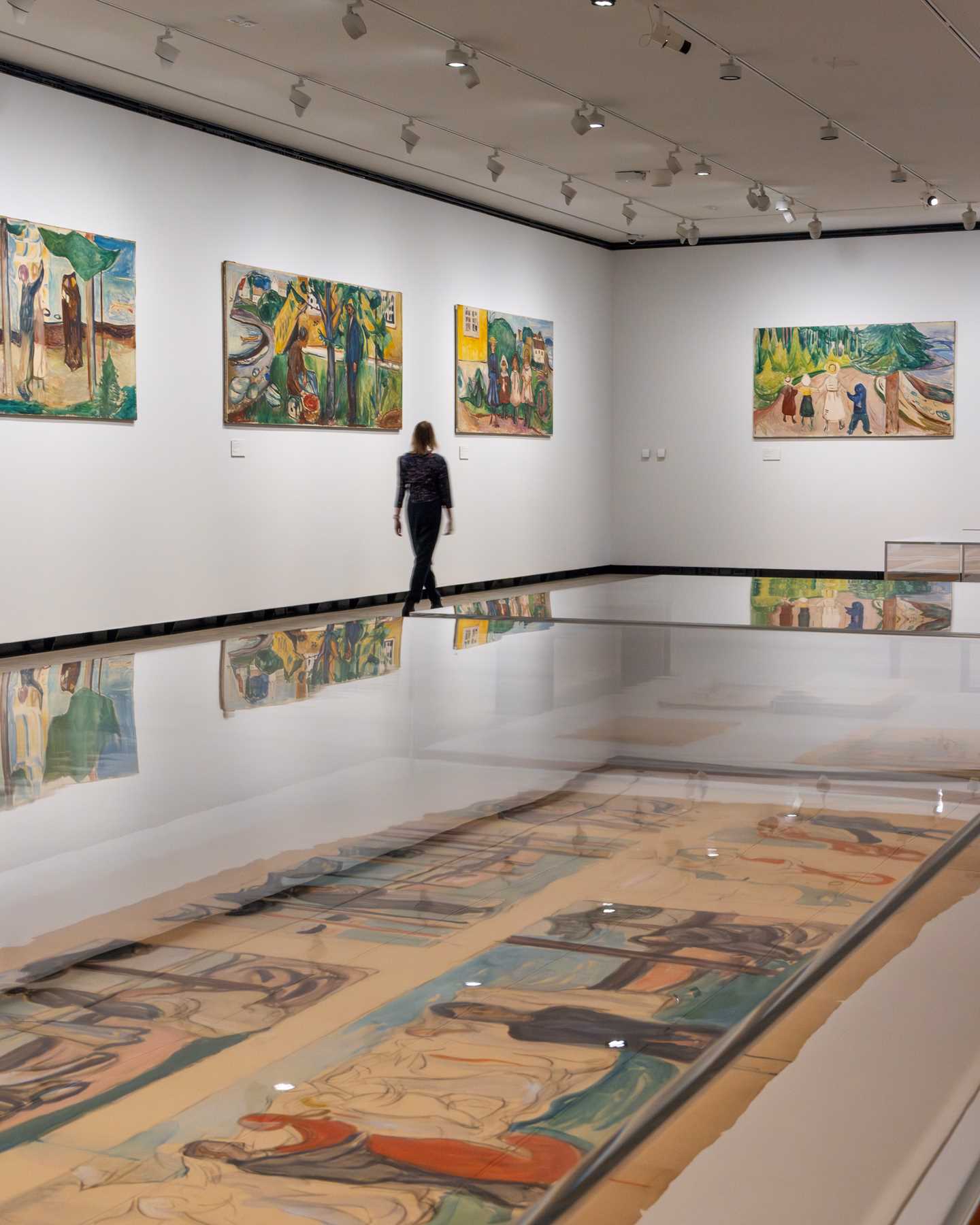 In the foreground, a glass display case contains sketches. In the background, a woman walks along a white wall, looking at the paintings hanging there. Photograph.