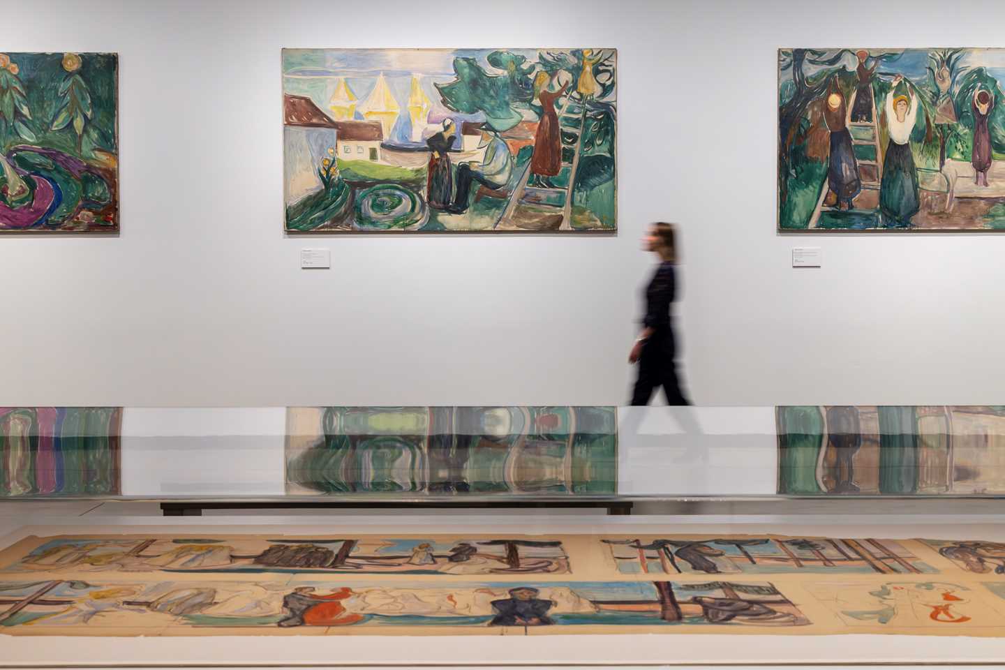 In the foreground is a glass display case containing sketches. In the background, a woman walks along a white wall while looking at the paintings hanging there. Photograph.