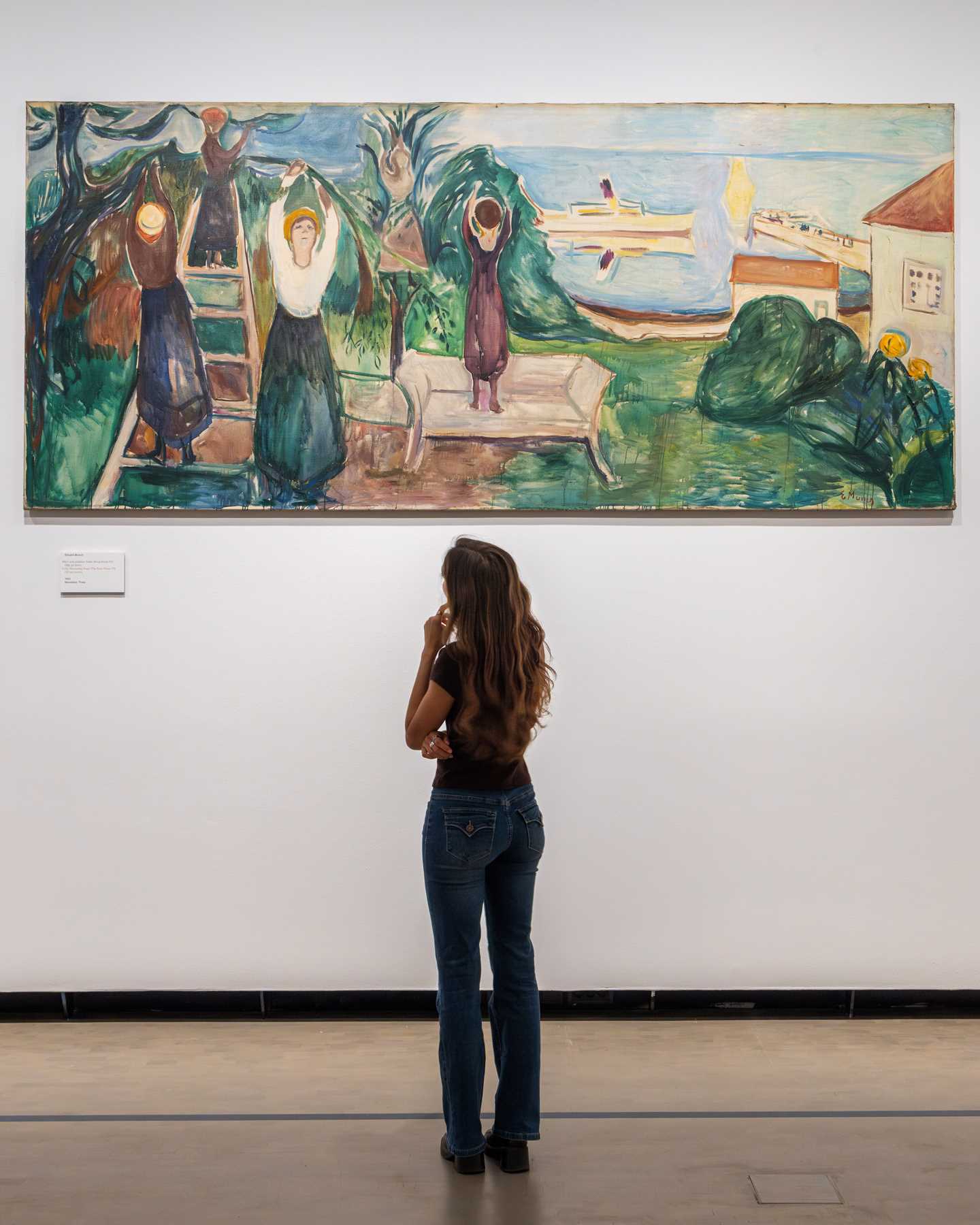 A woman stands with her back to the camera, looking up at a work from the Freia Frieze hanging on the wall. The painting depicts women picking fruit from trees, with the sea and a boat either docked or passing by in the background. Photograph.