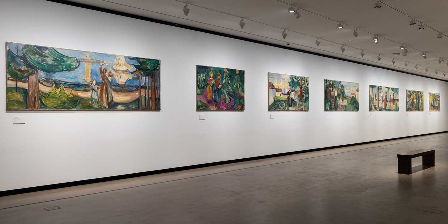 A long white wall in an exhibition gallery displaying seven of the twelve works in the Freia Frieze by Edvard Munch. The paintings depict summer scenes from Åsgårdstrand. Photograph.