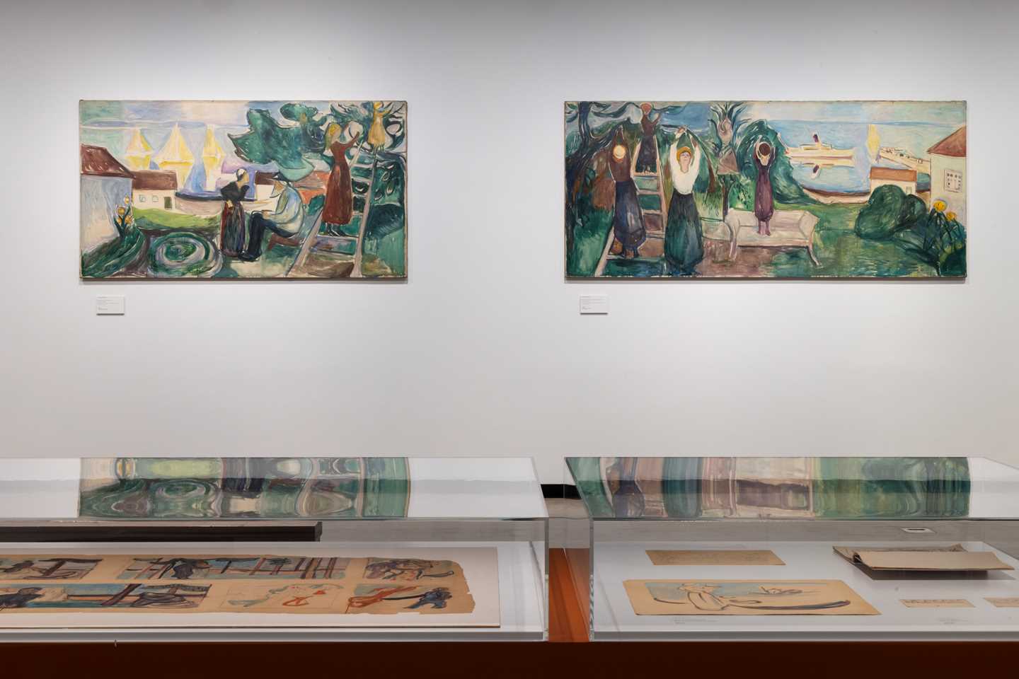 A white wall where two paintings are hanging. In the foreground, a glass display case contains sketches for the paintings shown on the wall. Photograph.