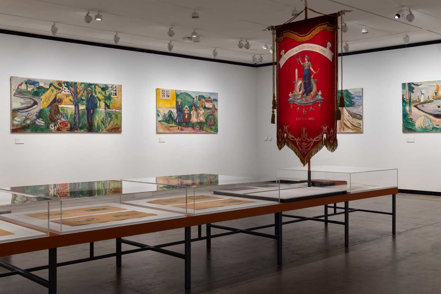 Two white walls meet in a corner, each displaying works from the Freia Frieze. In the foreground, a glass display case presents sketches for the Freia Frieze, along with an original banner from the Freia chocolate factory. Photograph.