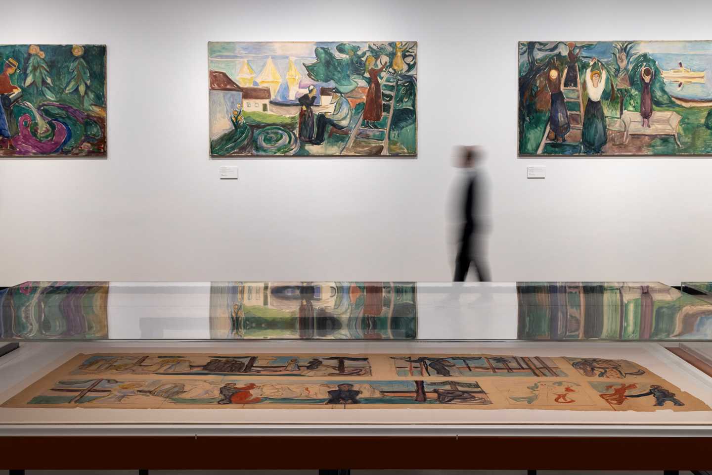 A man moves along a wall where three paintings are hanging. In the foreground, a glass display case contains sketches for the paintings shown on the wall. Photograph.