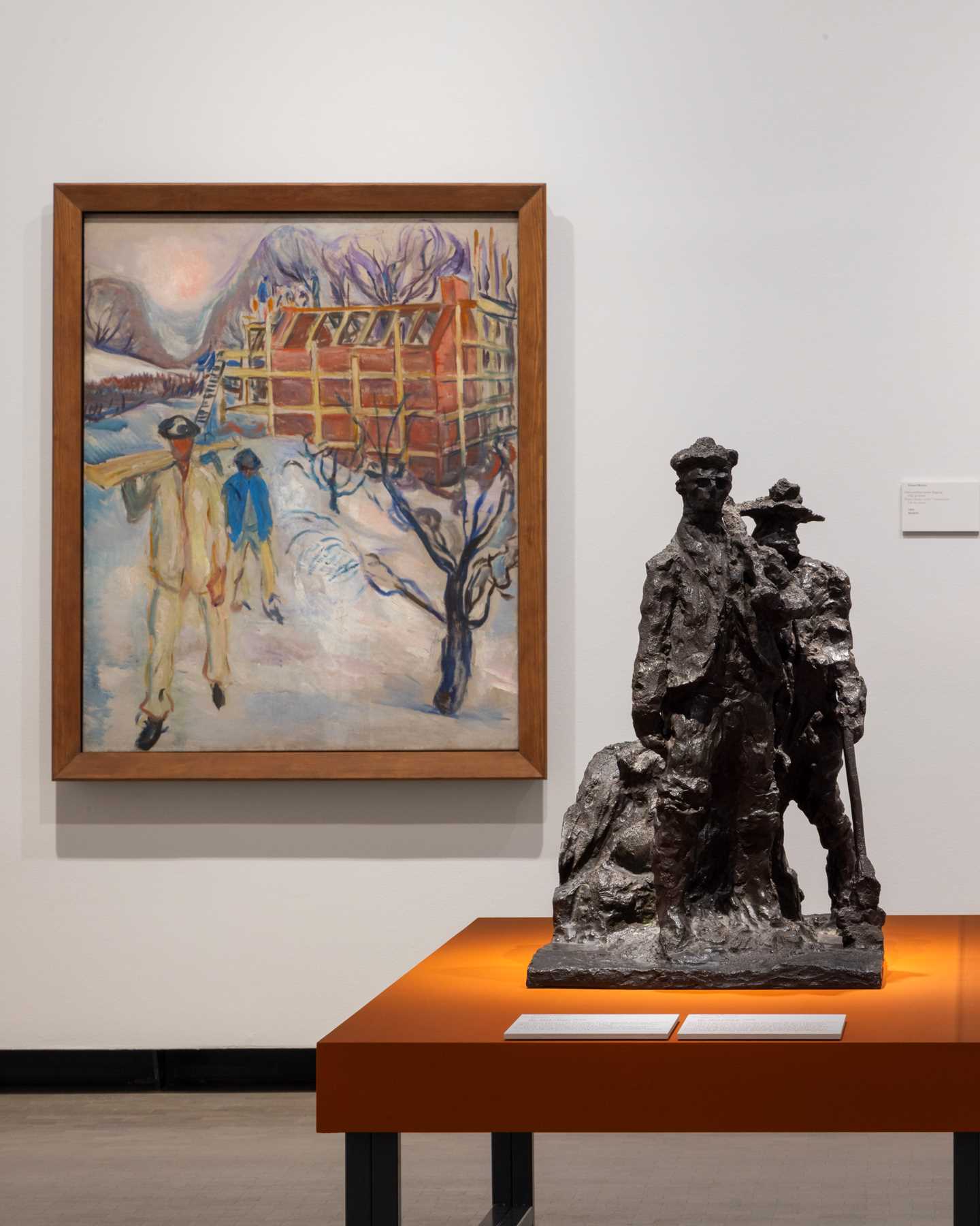 A bronze sculpture of workers stands on a table in the foreground of the image. On the wall behind the sculpture hangs a painting of a winter landscape with two workers building a house. Photograph.