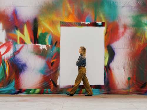 Artist Katharina Grosse walking in profile from right to left, with a wall-sized artwork by Grosse behind her. Photograph.